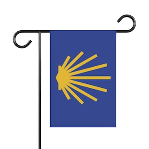 May include: A blue garden flag with a yellow scallop shell emblem. The flag is displayed on a black metal stand. The shell design has a central point with radiating lines. The flag is suitable for outdoor use.