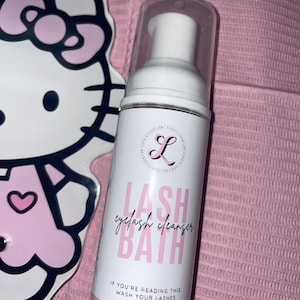 K&ouml;nnte beinhalten: Eine wei&szlig;e und rosa Wimpernreinigerflasche mit dem Text "LASH eyelash cleanser BATH" und "IF YOU'RE READING THIS, WASH YOUR LASHES." Im Hintergrund befindet sich ein Hello Kitty-f&ouml;rmiger Spiegel. Die Flasche enth&auml;lt 60 ml.
