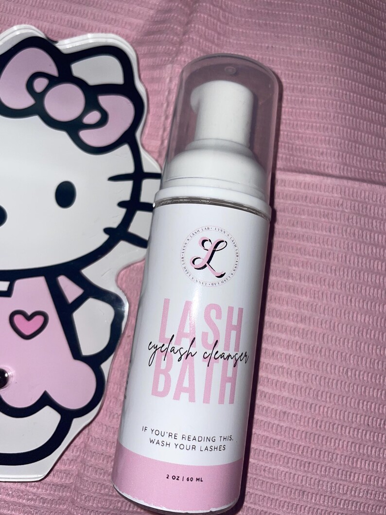K&ouml;nnte beinhalten: Eine wei&szlig;e und rosa Wimpernreinigerflasche mit dem Text "LASH eyelash cleanser BATH" und "IF YOU'RE READING THIS, WASH YOUR LASHES." Im Hintergrund befindet sich ein Hello Kitty-f&ouml;rmiger Spiegel. Die Flasche enth&auml;lt 60 ml.
