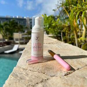 K&ouml;nnte beinhalten: Eine wei&szlig;e Flasche Wimpernreiniger, ein rosa-goldener Applikator und ein rosa Wimpernkamm. Auf der Flasche steht "Lash Bath". Die Artikel befinden sich auf einer Steinoberfl&auml;che mit einem Pool und tropischen Pflanzen im Hintergrund.