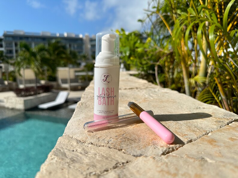 K&ouml;nnte beinhalten: Eine wei&szlig;e Flasche Wimpernreiniger, ein rosa-goldener Applikator und ein rosa Wimpernkamm. Auf der Flasche steht "Lash Bath". Die Artikel befinden sich auf einer Steinoberfl&auml;che mit einem Pool und tropischen Pflanzen im Hintergrund.