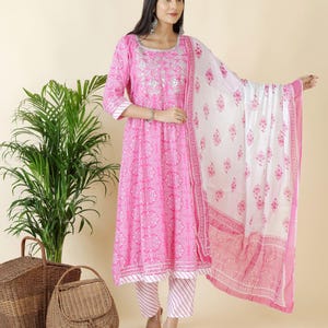 Puede incluir: Un vibrante conjunto de salwar kameez indio rosa. El conjunto incluye una túnica larga con intrincados bordados blancos, pantalones a juego con un patrón de rayas y una dupatta blanca con diseños florales rosas. Estilo de ropa tradicional.