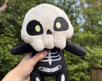 May include: A plush skeleton doll with a white skull and black body. The doll has black arms and legs, and a white rib cage design. The eyes are large, black circles with small, light green centers. The doll is soft and cuddly.
