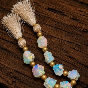 May include: Two strands of jewelry featuring raw, iridescent opal beads interspersed with smooth, matte gold-toned metal spheres. Each strand is finished at the top with a beige silk tassel secured by a matching gold-toned metal cap.
