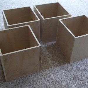 May include: Four unfinished wooden storage boxes. Each box is a cube shape with open tops and light brown wood grain. The boxes are arranged on a light-colored carpet.