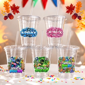 Può includere: Cinque bicchieri in plastica trasparente con coperchi e cannucce. Due bicchieri superiori presentano loghi Roblox in blu e rosa. Tre bicchieri inferiori mostrano grafiche colorate Roblox Brain Rot con vari personaggi e disegni.