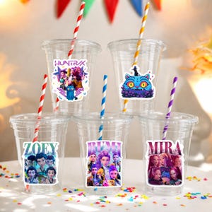 May include: Clear plastic cups with colorful stickers and striped straws. The stickers feature cartoon characters and the names "HUNTRAX", "ZOEY", "RUMI", and "MIRA". The straws are red and white, blue and white, yellow and white, and purple and white.