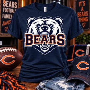 Puede incluir: Una camiseta de manga corta azul marino con un gráfico grande de una cara de oso blanco gruñendo y la palabra BEARS impresa en letras mayúsculas de color naranja y blanco en el centro. La camiseta tiene cuello redondo y un corte estándar.