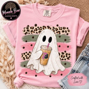May include: A light pink short-sleeve t-shirt featuring a central graphic of a white ghost holding an iced coffee cup with the number 73 on it. The design includes horizontal brush strokes in leopard print, sage green, and pink with black dots.