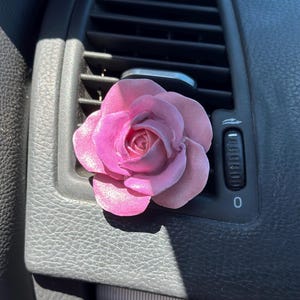 Könnte beinhalten: Ein rosa Rosen-Lufterfrischer, der an der Lüftung eines Autos hängt.