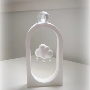 Puede incluir: Escultura de cerámica blanca de una nube con gotas de lluvia colgando. La escultura está dentro de un marco blanco con una bola de cristal transparente en la parte superior. La escultura está sobre una superficie blanca.