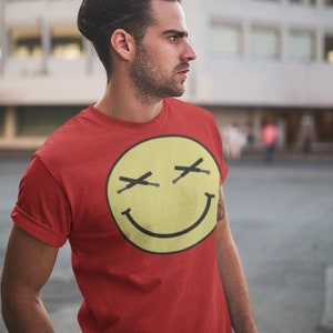 Pode incluir: Um homem vestindo uma camiseta vermelha com um gráfico de um rosto sorridente amarelo. O rosto sorridente tem X's pretas para os olhos.