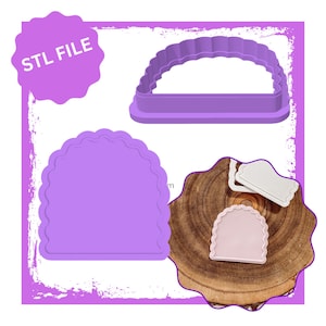 Op de afbeelding: Paarse plastic koekjesvormer in de vorm van een halve cirkel met gegolfde randen. De vormer wordt getoond op een houten oppervlak met twee kleinere vormen in dezelfde vorm.