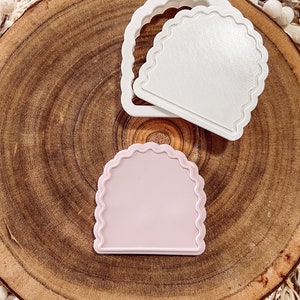May include: Two plastic cookie cutters in the shape of an arch with scalloped edges. One cutter is white and the other is light pink.