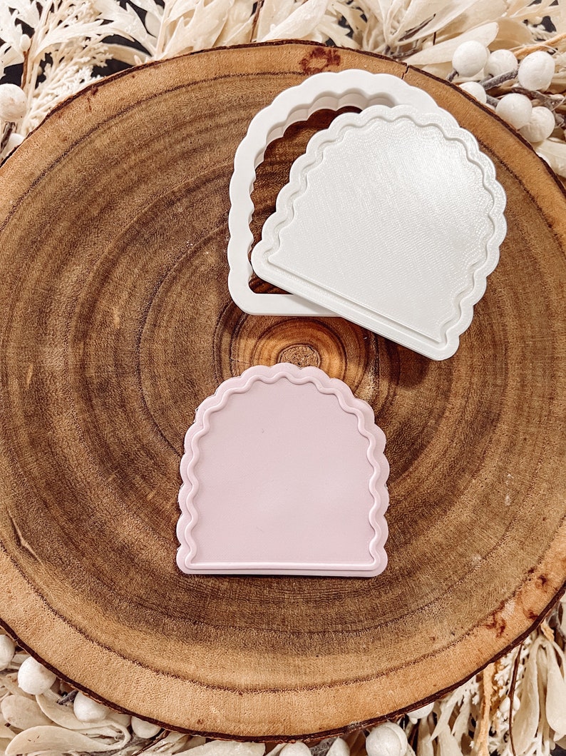 May include: Two plastic cookie cutters in the shape of an arch with scalloped edges. One cutter is white and the other is light pink.