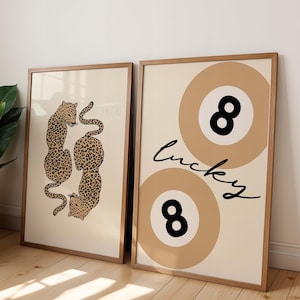 May include: Two framed prints with a beige background. The print on the left features a pair of leopards in a curled position. The print on the right features two beige circles with a black 8 inside each circle. The text "lucky" is written in cursive on the right print.