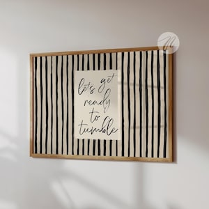 May include: A rectangular wall art piece in a light wood frame featuring a beige background with vertical black stripes. Centered is a white rectangular panel with the black handwritten text "lets get ready to tumble" in a cursive script font.