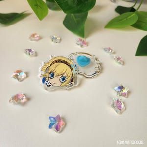 May include: A clear acrylic pin featuring a cartoon character with blonde hair and blue eyes. The pin is surrounded by iridescent star-shaped crystals and a small, heart-shaped pin. Green leaves are in the background.