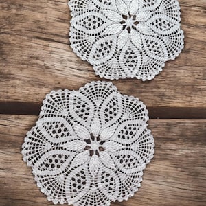 May include: Two white crocheted doilies with a floral design on a wooden surface.