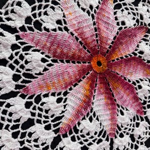 May include: White crochet doily with a pink and orange flower in the center.