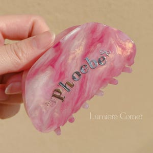 May include: Pink and white marbled hair claw clip with the name "phoebe" in silver lettering. The clip has a curved shape and a claw design for securing hair. The text "Lumiere Corner" is also visible.