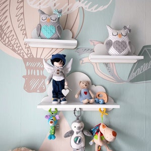 Op de afbeelding: Drie witte wolkvormige planken met een witte ster, een grijze uil, een pop met vleugels, een teddybeer en drie hangende speelgoedjes. De planken zijn gemonteerd op een lichtblauwe muur met een luchtballonpatroon.