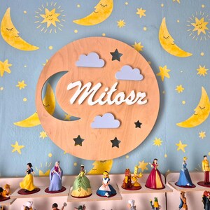 Può includere: Un cartello di legno a forma di luna con il nome "Mitosz" scritto in lettere bianche. Il cartello è decorato con nuvole bianche e stelle. Il cartello è appeso a un muro con un motivo di luna e stelle blu e giallo.