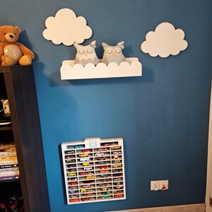 May include: A white wooden shelf with a scalloped edge, shaped like a cloud, with two gray owl stuffed toys on top. The shelf is mounted on a blue wall with two white wooden clouds on either side. Below the shelf is a white wooden toy car storage rack with the letter "P" at the top, filled with miniature toy cars.