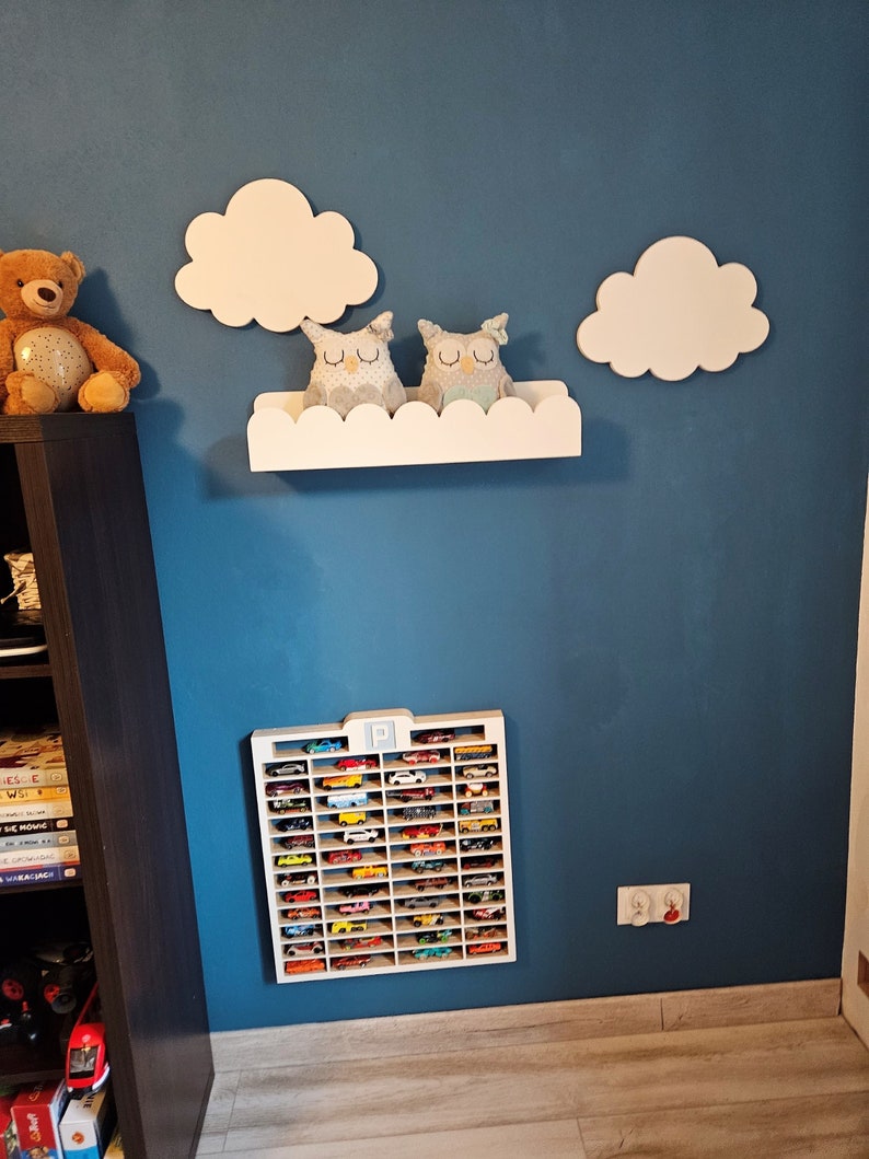 May include: A white wooden shelf with a scalloped edge, shaped like a cloud, with two gray owl stuffed toys on top. The shelf is mounted on a blue wall with two white wooden clouds on either side. Below the shelf is a white wooden toy car storage rack with the letter "P" at the top, filled with miniature toy cars.