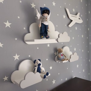 K&ouml;nnte beinhalten: Drei wei&szlig;e, wolkenf&ouml;rmige Regale vor einem grauen Hintergrund. Das obere Regal hat eine Puppe mit blauen Fl&uuml;geln und einem blau-wei&szlig;en Pullover. Das mittlere Regal hat einen Teddyb&auml;r mit einem roten Herzen. Das untere Regal ist leer.