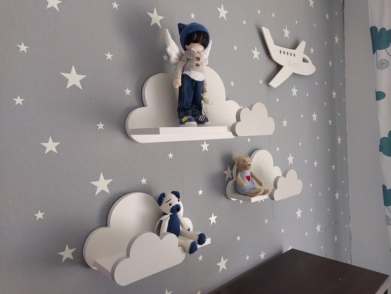 K&ouml;nnte beinhalten: Drei wei&szlig;e, wolkenf&ouml;rmige Regale vor einem grauen Hintergrund. Das obere Regal hat eine Puppe mit blauen Fl&uuml;geln und einem blau-wei&szlig;en Pullover. Das mittlere Regal hat einen Teddyb&auml;r mit einem roten Herzen. Das untere Regal ist leer.