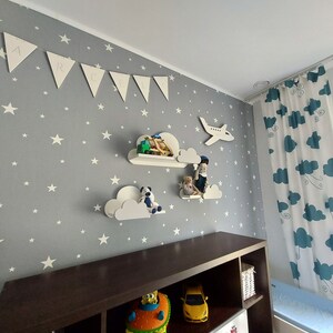 K&ouml;nnte beinhalten: Ein Kinderzimmer mit einer grauen Wand, die mit wei&szlig;en Sternen bedeckt ist. Es gibt wei&szlig;e, wolkenf&ouml;rmige Regale mit Spielzeug darauf. Ein Holzregal mit Spielzeug und ein wei&szlig;er Teddyb&auml;r mit dem Wort "Marcel" darauf. Ein wei&szlig;er Flugzeug-Ausschnitt ist an der Wand. Ein wei&szlig;es Banner mit den Buchstaben "MARCEL" ist an der Wand. Ein wei&szlig;er und blauer Vorhang mit einem Wolkenmuster ist auf der rechten Seite des Bildes.