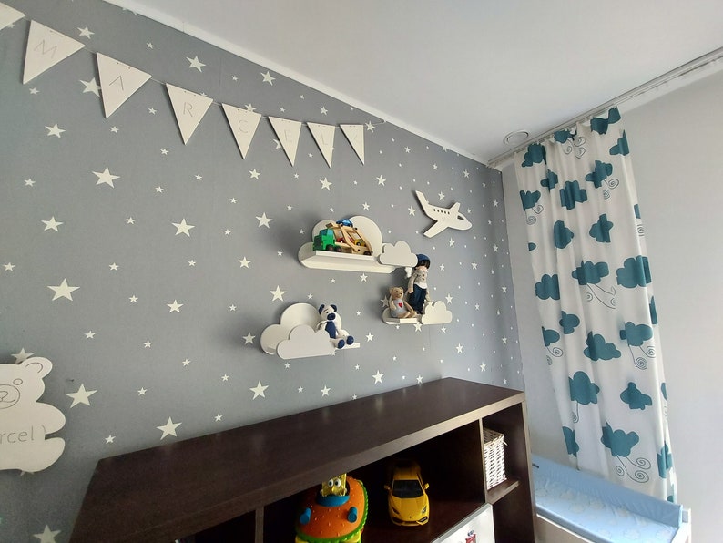 K&ouml;nnte beinhalten: Ein Kinderzimmer mit einer grauen Wand, die mit wei&szlig;en Sternen bedeckt ist. Es gibt wei&szlig;e, wolkenf&ouml;rmige Regale mit Spielzeug darauf. Ein Holzregal mit Spielzeug und ein wei&szlig;er Teddyb&auml;r mit dem Wort "Marcel" darauf. Ein wei&szlig;er Flugzeug-Ausschnitt ist an der Wand. Ein wei&szlig;es Banner mit den Buchstaben "MARCEL" ist an der Wand. Ein wei&szlig;er und blauer Vorhang mit einem Wolkenmuster ist auf der rechten Seite des Bildes.