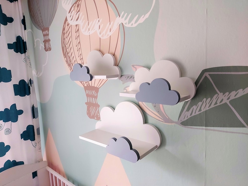 K&ouml;nnte beinhalten: Drei wei&szlig;e Wolkenf&ouml;rmige Regale mit grauen Wolkenakzenten, die an einer hellgr&uuml;nen Wand mit einem Hei&szlig;luftballonmuster montiert sind.