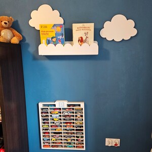 May include: A white shelf with a cloud design holds two children's books, one titled "Audiobajki Magia Słuchania" and the other titled "Moje pierwsze literki".  A white cloud-shaped wall decor is on the wall above the shelf.  A white toy car garage with a black "P" is on the wall below the shelf.