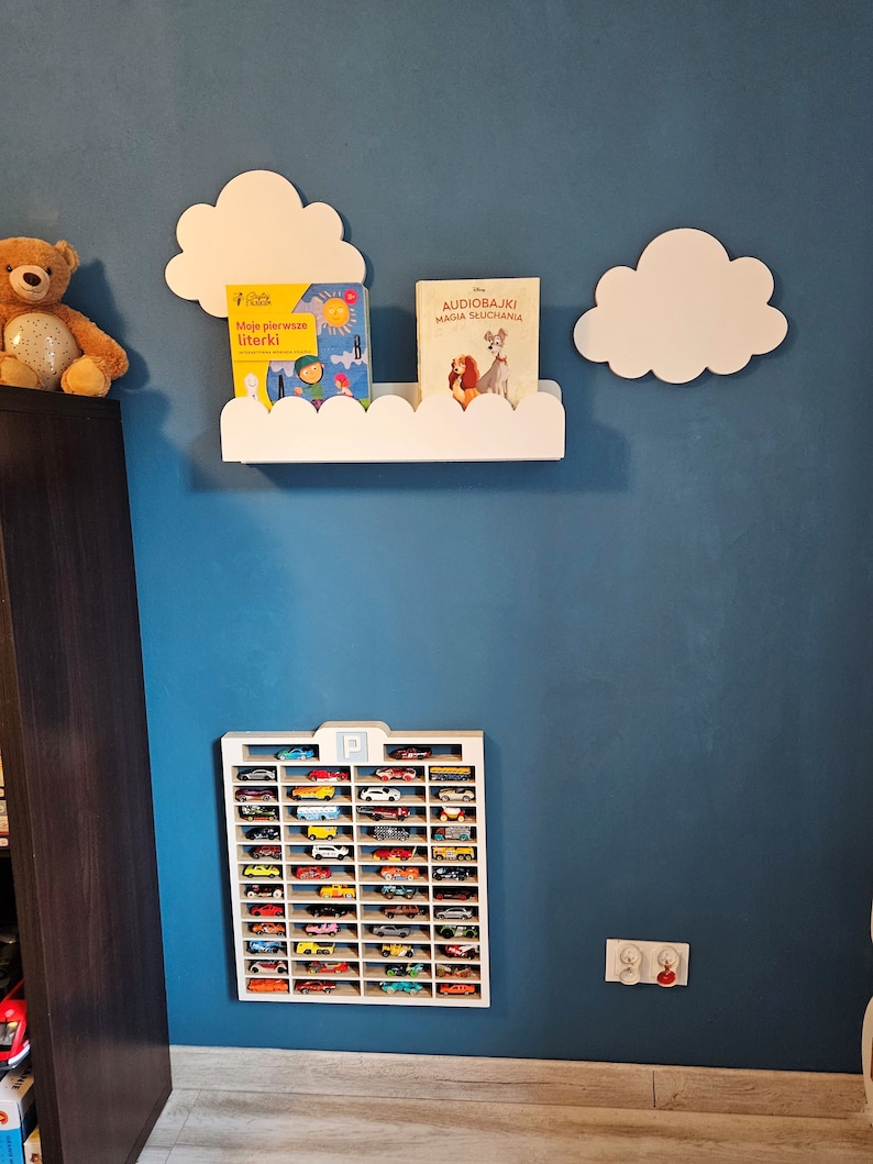 May include: A white shelf with a cloud design holds two children's books, one titled "Audiobajki Magia Słuchania" and the other titled "Moje pierwsze literki".  A white cloud-shaped wall decor is on the wall above the shelf.  A white toy car garage with a black "P" is on the wall below the shelf.