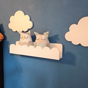 May include: White cloud-shaped shelves with scalloped edges, mounted on a blue wall. Two plush owl toys are sitting on the shelves. A brown teddy bear is on a shelf below.