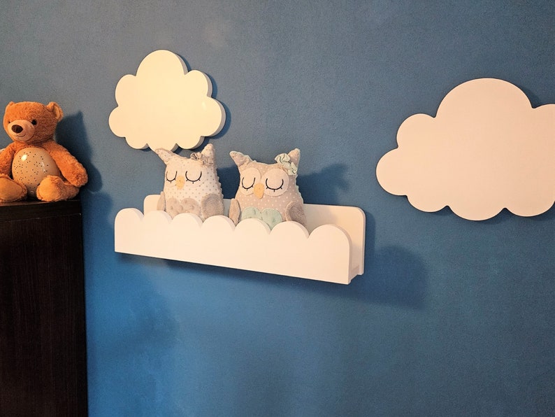 May include: White cloud-shaped shelves with scalloped edges, mounted on a blue wall. Two plush owl toys are sitting on the shelves. A brown teddy bear is on a shelf below.