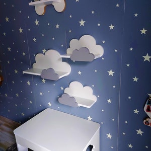 K&ouml;nnte beinhalten: Wei&szlig;er Holztisch und Stuhl-Set mit einer blauen Wand, die mit wei&szlig;en Sternen und wolkenf&ouml;rmigen Regalen verziert ist. Die Regale sind grau und wei&szlig; lackiert.