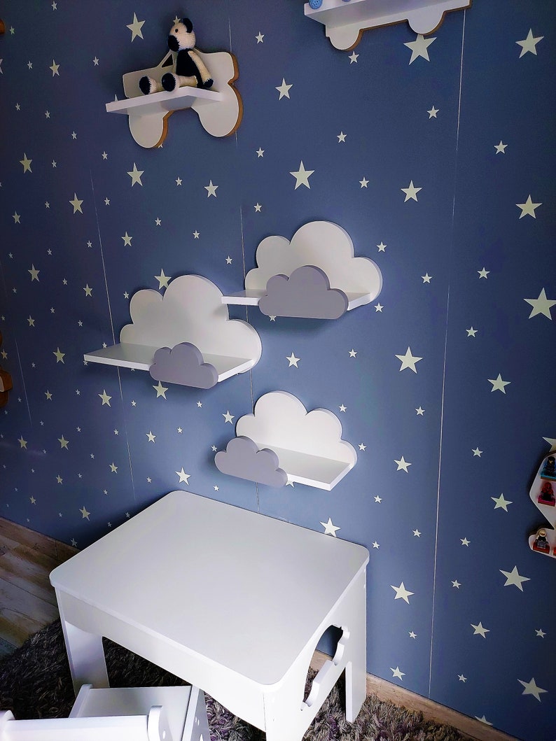 K&ouml;nnte beinhalten: Wei&szlig;er Holztisch und Stuhl-Set mit einer blauen Wand, die mit wei&szlig;en Sternen und wolkenf&ouml;rmigen Regalen verziert ist. Die Regale sind grau und wei&szlig; lackiert.