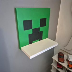 Op de afbeelding: Een groen Minecraft Creeper gezicht wandkunst met een wit plankje bevestigd. Het Creeper gezicht is gemaakt van groene en zwarte vierkanten. Het plankje is rechthoekig en wit geschilderd.