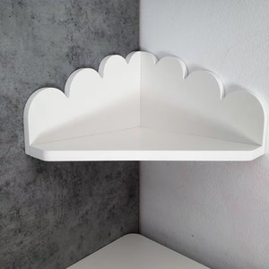 Könnte beinhalten: Weißes, wolkenförmiges Eckregal. Das Regal ist für eine Ecke konzipiert und hat eine flache Oberfläche zum Präsentieren von Gegenständen. Das Regal ist aus Holz gefertigt und weiß lackiert. Es ist an einer grau-weißen Wand montiert.