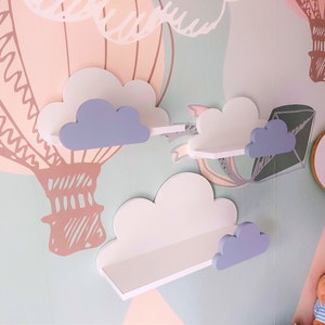 K&ouml;nnte beinhalten: Drei wei&szlig;e, wolkenf&ouml;rmige Regale mit hellblauen Wolkenakzenten, die an einer hellblauen Wand montiert sind. Die Regale sind mit einem Hei&szlig;luftballon-Motiv verziert.