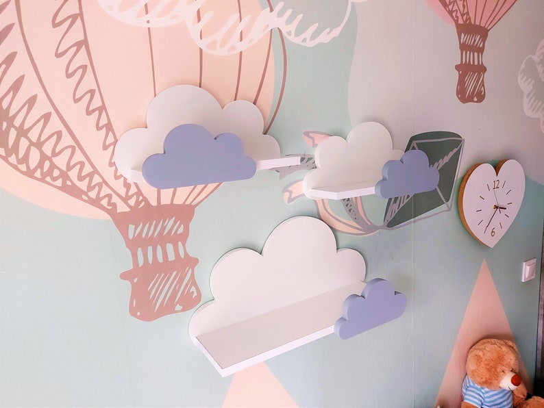 K&ouml;nnte beinhalten: Drei wei&szlig;e, wolkenf&ouml;rmige Regale mit hellblauen Wolkenakzenten, die an einer hellblauen Wand montiert sind. Die Regale sind mit einem Hei&szlig;luftballon-Motiv verziert.