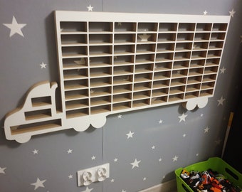 Puede incluir: Estantería blanca de madera con forma de camión con numerosos compartimentos pequeños, montada en una pared gris decorada con pegatinas de estrellas blancas. La estantería está diseñada para guardar coches de juguete pequeños.