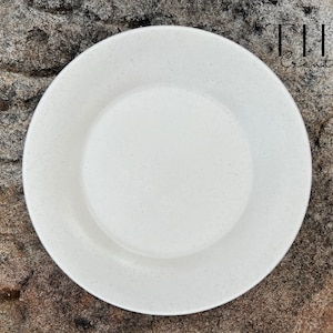 May include: A white speckled ceramic plate with a wide rim and a recessed centre. The plate is set against a textured, neutral-toned background. The text "THIO Creations" is visible in the upper right corner.