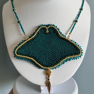 May include: A handmade necklace featuring a large, emerald green beaded pendant with a gold-colored border. The pendant is suspended from a beaded chain. A gold-colored spiral and arrowhead charm dangles from the pendant.