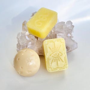 May include: Three yellow soap bars with different designs. One bar has a star pattern, one has an eye design, and one has the word "GOOD" embossed on it. The soap bars are sitting on a bed of white crystals.