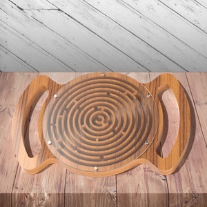 May include: A wooden circular maze with a clear plastic cover. The maze has a handle on each side. The maze is on a wooden surface.
