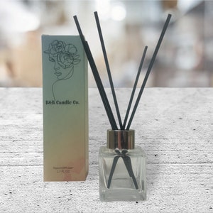 Puede incluir: Una botella de vidrio transparente con tapa dorada y cañas negras, etiquetada "B&B Candle Co. Reed Diffuser 1.7 FL OZ".